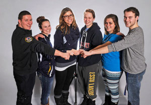 six students standing in front of a gray background putting their hands together
