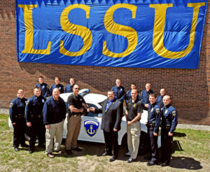 MCOLES Academy students, instructors and the Chippewa County Sheriffs Department shaking hands for the department donating a partrol car to the Acadmey, all under a large blue and gold LSSU flag