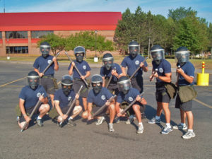 10 MCOLES students standing in front on the Norris building all wearing riot gear on a sunny day