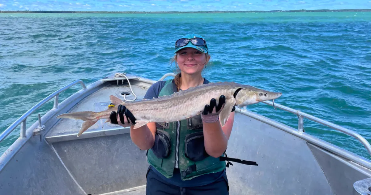 Student holding a sturgeon on a fishing boat out on the water.