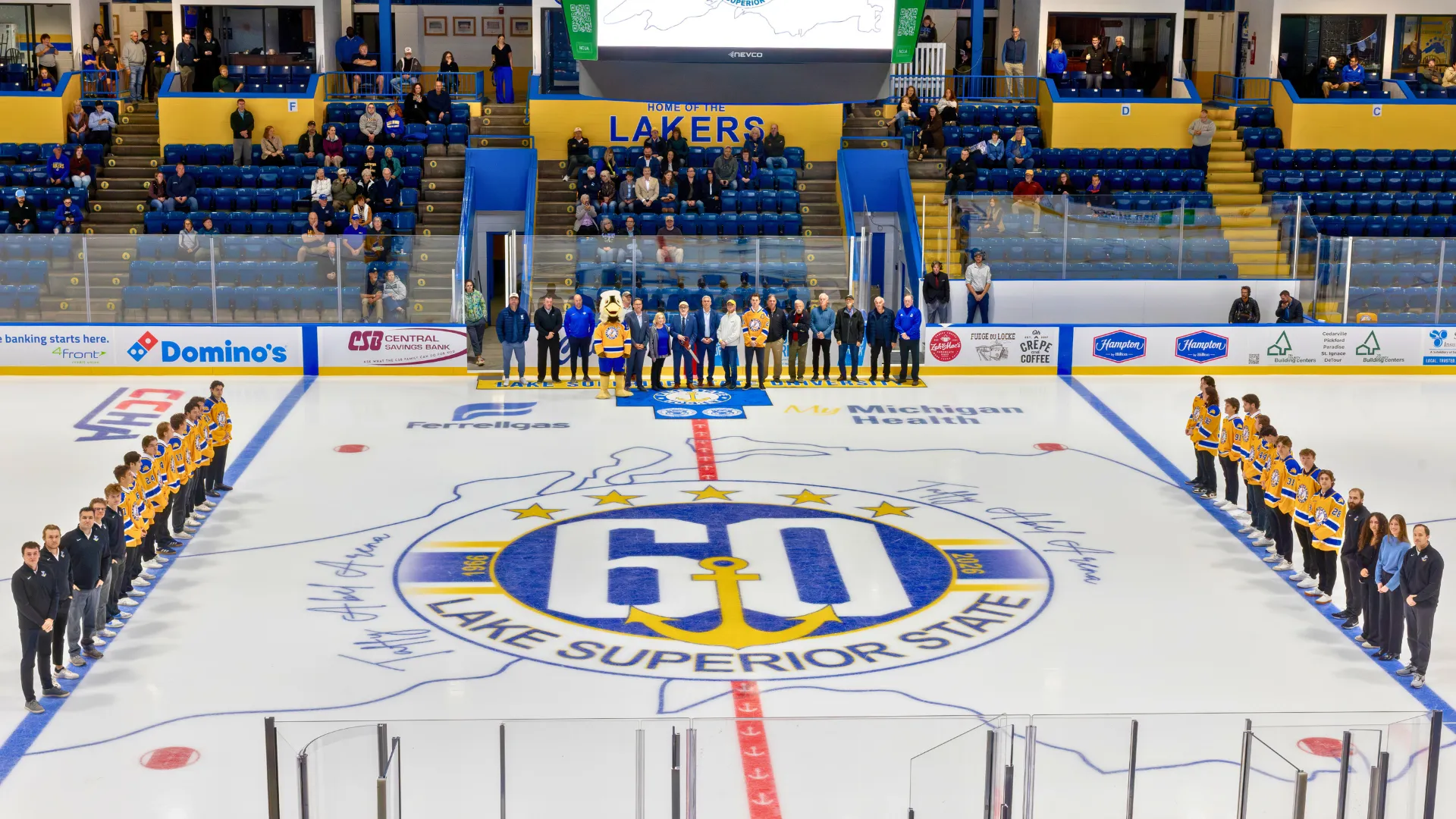 Arial view of LSSU hockey rink, team member lined up along both blue lines, and fans in the stands and along the boards on the far side of the rink.