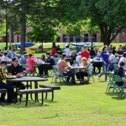 Orientation attendees seating at tables eating lunch with campus buildings in the background.
