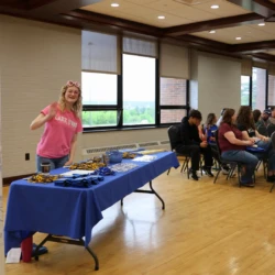 Admissions representative waving at camera behind table of LSSU merchandise with some seated attendees visible.