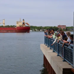 Freighter entering frame while community members are gathered alongside the barrier, overlooking Atlantic Salmon as the enter the St. Mary's river during the release event.