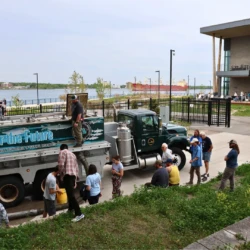 MDNR release truck is surrounding by onlookers as they observe Atlantic Salmon travel down the attached tube to the release drain. A freighter and CFRE is visible in the background.