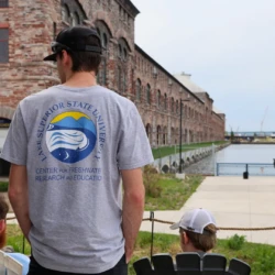 The back of a student's shirt featuring the CFRE logo is highlighted, with community members and the power plant that houses CFRE's hatchery in the background.
