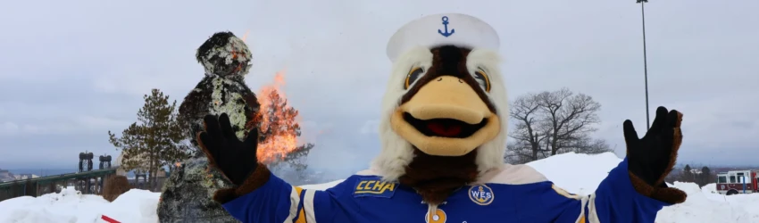 Seamore the Sea Duck raises his hands, wearing an LSSU jersey, in front of a paper snowman actively on fire at the annual snowman burn event.