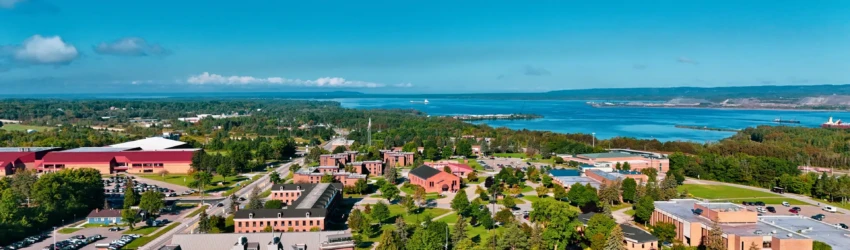 Drone image of LSSU campus and the surrounding greenery, including a freighter visible on the water in the background, all under clear blue skies.