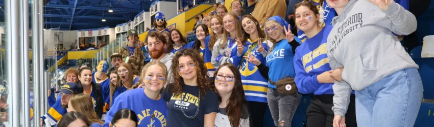 LSSU students in the student section at a Laker hockey game, wearing LSSU attire, many with face paint, all facing the camera and smiling.