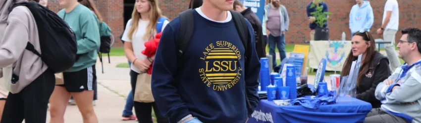 LSSU Student walking on campus, wearing an LSSU vintage design hoodie, with other students, tables, and organization representatives visible at the annual Lakerpalooza event.