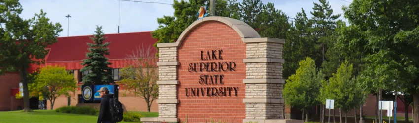 A students walking past a brick sign with a rounded top that reads 'Lake Superior State University'.