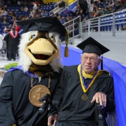 Seamore the Sea Duck kneels alongside the eldest golden grad in attendance.