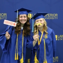 Graduates in caps and gowns with photo booth accessory pieces in front of a LSSU backdrop.