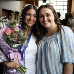 Graduate and supporter smile at camera, pinning recipient holding a bouquet of flowers.
