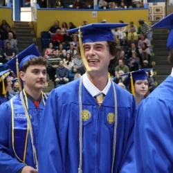 Graduates in caps and gowns enter the arena for commencement. One graduate is seen with a candid smile looking at supporters in the crowd.