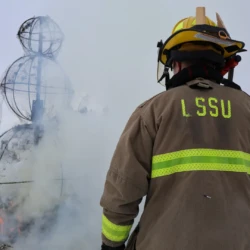 LSSU Fire Science student in gear onlooks snowman metal remains after fire is extinguished.