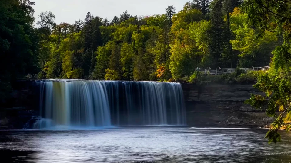 Tahquamenon falls with rushing water falling over a rocky cliff surrounded by trees. A few onlookers can be seen against a railing.