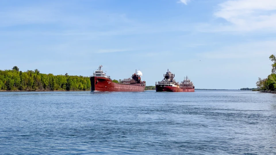 Two red freighters side-by-side in water with green trees visible behind them.